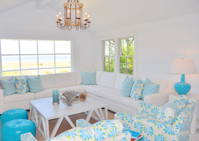 Living room in a cottage with turquoise pillows on a white sectional, two turquoise moroccan poufs, turquoise and white armchairs, a sea grass rug, white coffee table and a chandelier. 