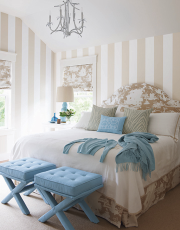 bedroom with upholstered headboard, blue ottomans at the foot of the bed, striped beige and white walls and a chandelier
