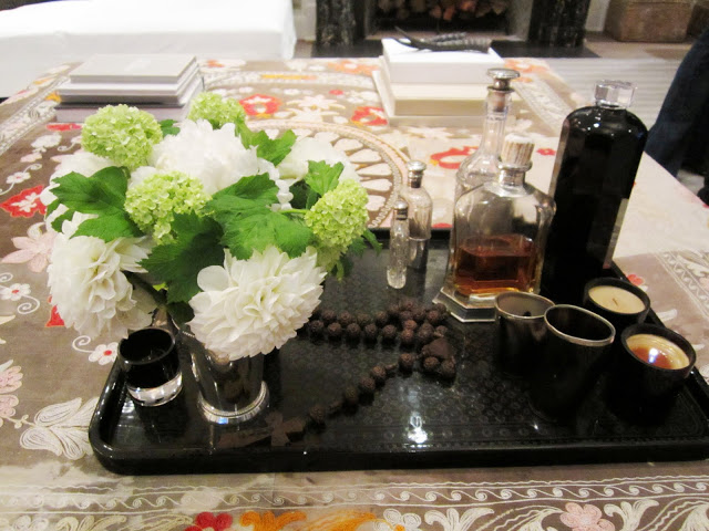 Close up of a black tray on the patterned ottoman. On the tray are prayer beads, candles, assorted bottles and a cup full of white hydrangeas