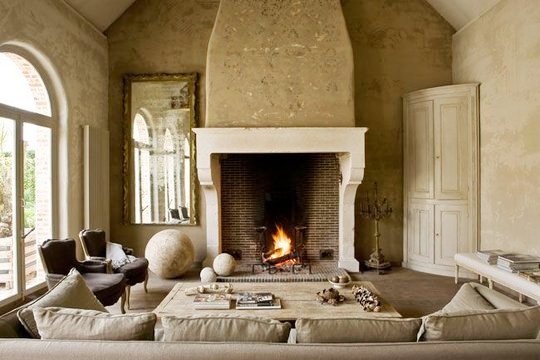 Mansion in Belgium with barn converted into a living room with a large fireplace with molded mantel, taupe sofa, light wood coffee table, two wingback chairs, a large traditional mirror and light wood cabinet