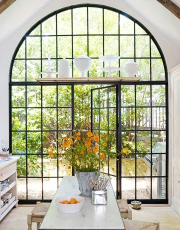 Kitchen with an large arched encasement window with black trim, floating shelves and a door through the "window" going outside to a lush garden