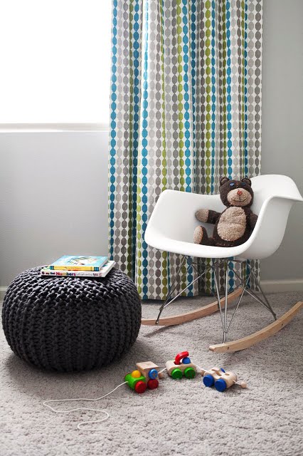 Kid's room with gray carpet, gray braided poof seat, floor length curtains with turquoise, green and gray spots in rows running down it, and a white Eames chair with a teddy bear in it