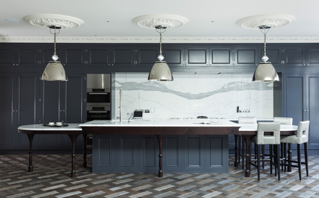 Alternative view of a kitchen in a suburban London home with blue grey cabinets, marble counter tops and backsplash, stainless appliances, silver pendant lights, carved crown moulding, ceiling medallions, parquet wood floors and leather barstools
