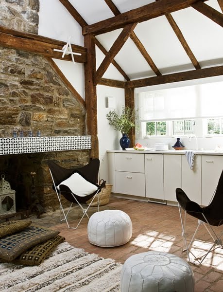 Inside a pool house with a brick floor, exposed wood beams, black butterfly chairs, white and grey Moroccan poufs, a stone fireplace, white cabinets and a striped shag rug. 