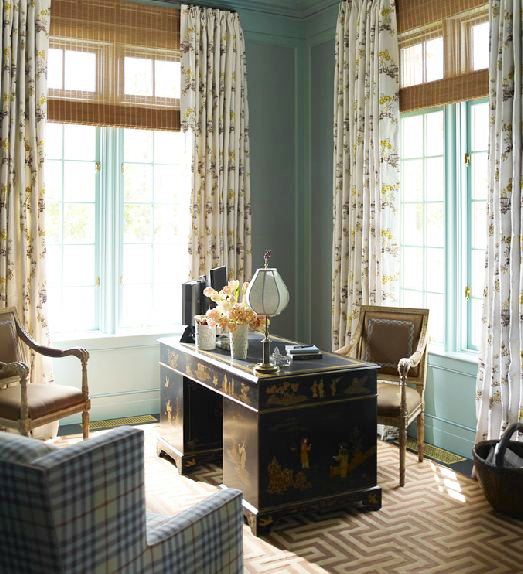 Library in Katie Ridder's home with a bold brown and cream graphic rug, rattan blinds, a plaid armchair across a Chinoisserie desk, blue walls with encasement windows covered in floor length floral curtains