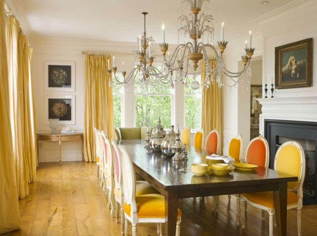 formal dining room in a san francisco mansion with orange and yellow upholstered louis XIV chairs, a dark wood farmhouse style table, wide paneled wood floor, floor length yellow curtains, two chrystal chandeliers and a fireplace with black molding and a white mantel