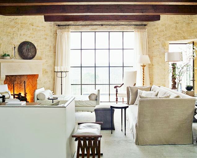 Living room with exposed beams, a large fireplace, a stone wall, large encasement window, taupe sofa and matching bench