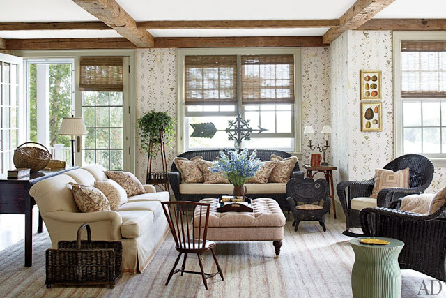 Living room in Nantucket with exposed beams, wood floors, floor to ceiling wall paper, french doors, wicker sofa and matching armchair with an upholstered sofa and an ottoman being used as a table. 