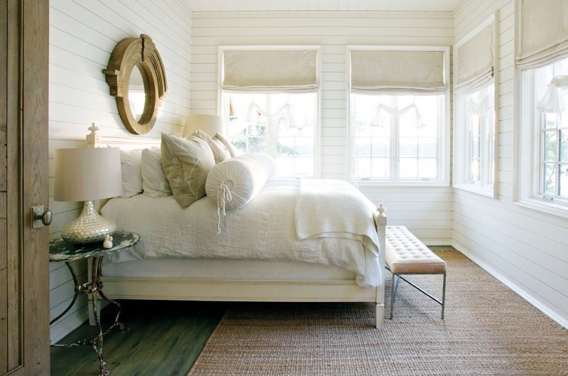 Rustic bedroom with white beadboard walls, a stained wood floor, sea grass rug, four windows with roman shades, an ottoman at the foot of the bed and a round mirror above the bed