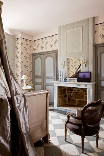 bedroom in a french mansion with pink and grey wallpaper, a painted checkered wood floor, a white fireplace, paneled doors and a Louis XIV chair