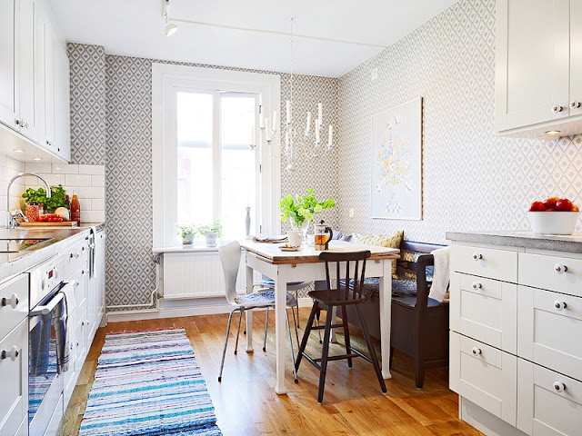 Alternative view of the kitchen in a tiny apartment with chandelier, geometric wallpaper, white cabinets and drawers, a cozy breakfast nook with a bench and mismatched chairs a wood floor and a striped rug