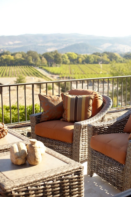 Patio with wicker armchairs with orange cushions and matching wicker coffe table overlooking a vineyard in Napa