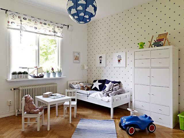 Child's room with one cream colored wall with blue poka dots, a white table and chair with a teddy bear in one of them, a toy car, a white bed with blue pillows, a white chest of drawers and a window with small plants on the window sill