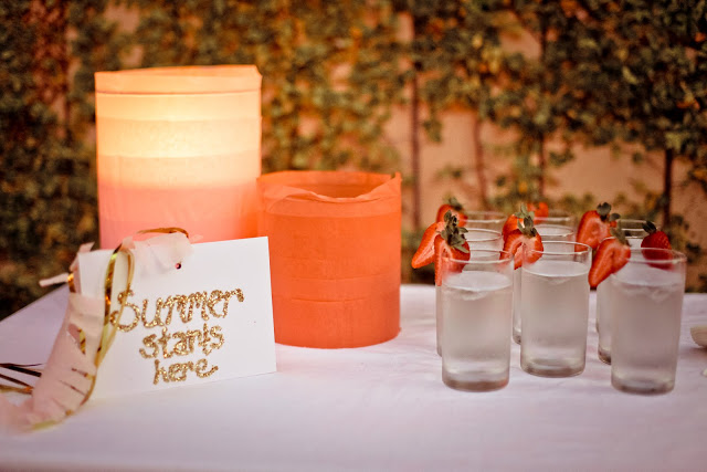 table with glasses of water with fresh strawberries on the rim, orange lanterns and a sign that says "Summer Starts Here"