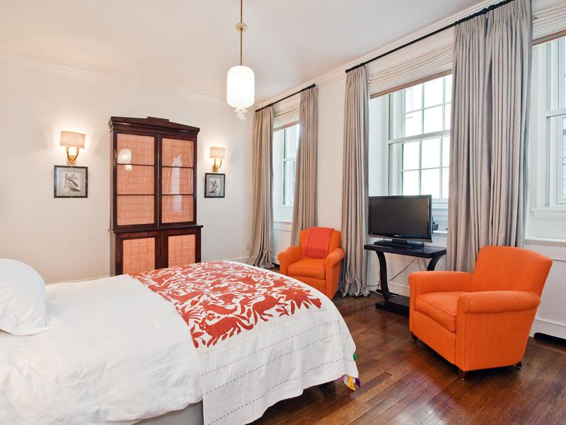 Guest bedroom in a park avenue apartment with orange club chairs, a television, wood floor, encasement windows, and an pendnat light