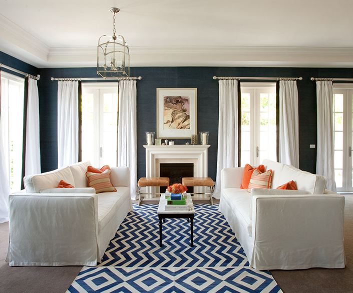 Living room with blue walls, dueling white sofas with orange accent pillows, and a blue and white geometric rug