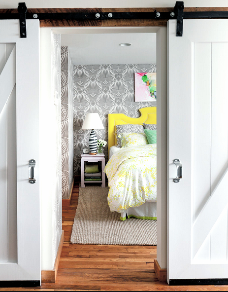 Barn doors open to reval a bedroom with gray Victorian inspired wallpaper, a white night stand, and a bed with yellow and white bedding and bright yellow molded headboard