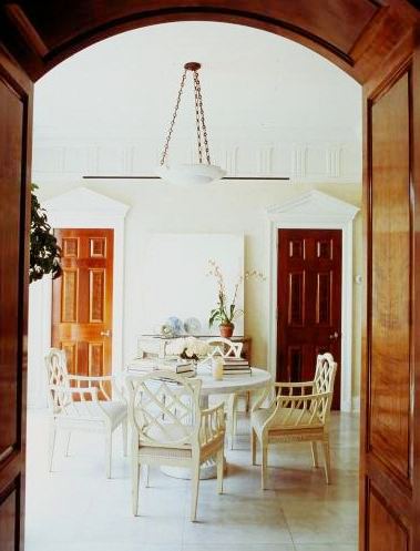Foyer by Michael S. Smith with white fretwork chairs around a white pedestal round table under a white pendant light, tile floor and two wood doors made of different kinds of wood