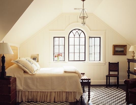 Attic bedroom with dark wood floor, a large black and white checkerboard rug, a pendant light, arched windows, a chest doubles as a nightstand, there is a carved bench at the foot of the bed with matching chair in the corner