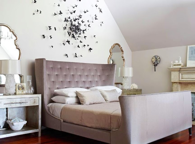 Bedroom with a fireplace and wood floors, pale purple tufted, wing back headboard, and two marbled nightstands on each side of the bed with silver leaf mirrors. Above the bed is a wall mounted sculpture made of butterflies