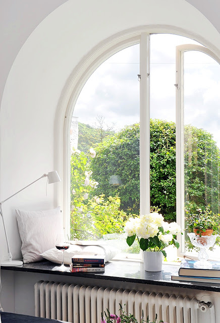 arched window opening to a beautiful view with window ledge holding a glass of wine, a stack of books, pillows, a reading lamp and a potted plant 