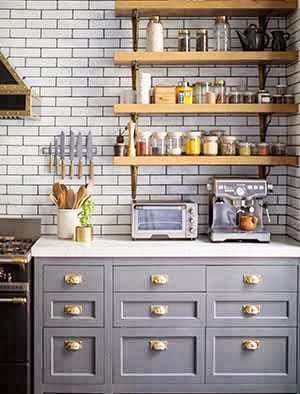Kitchen in a New York apartment with grey cabinets with brass pulls, wall mounted shelves, marble countertops and subway tile walls on dark grout