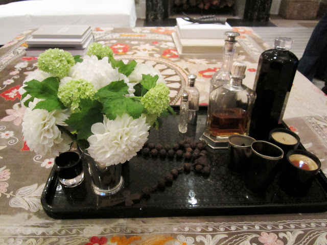  A large dark tray turns an ottoman into a coffee table vignette with green and white flowers, scented oils, a rosary, and candles