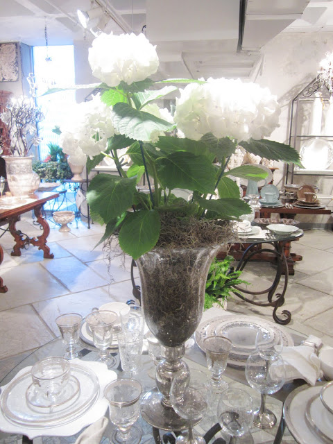 Close up of table setting and large white hydrangea plant in a glass vase on the round glass table above