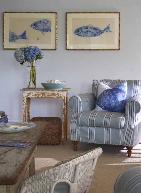 Living room with blue and white white striped armchair, reclaimed wood side table, white wicker chair and blue walls with framed pictures of blue fish