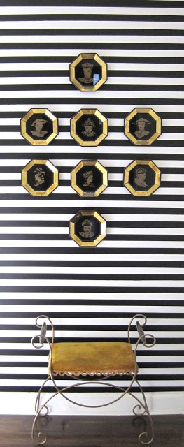 Hallway with black and white painted in horizontal stripes, 8 engraved portraits in gold octagon frames and an art deco metal stool with gold cushion