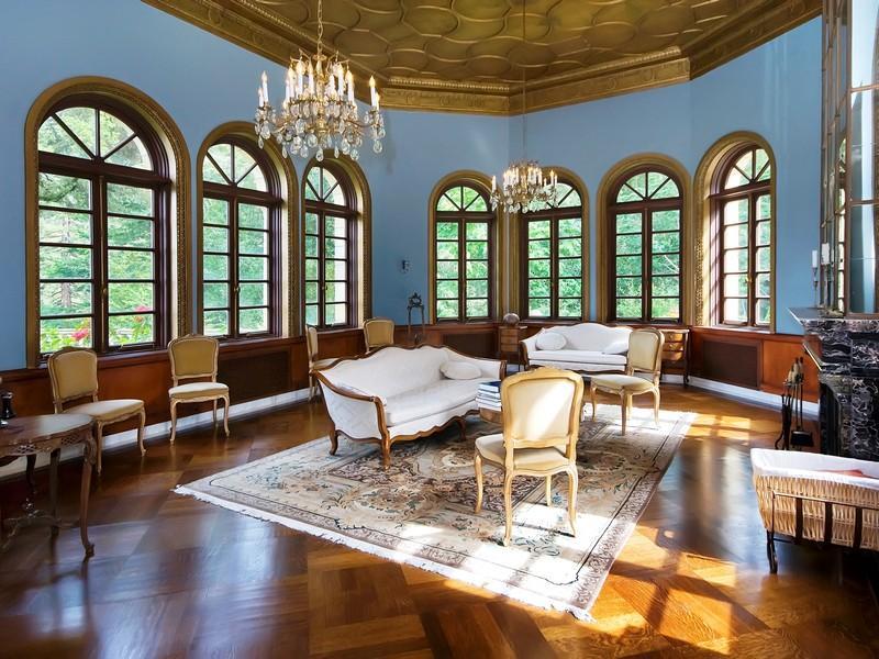 Living room with arched windows, crystal chandeliers, a coffered ceiling, wood floor, settees, a fireplace, and upholstered chairs