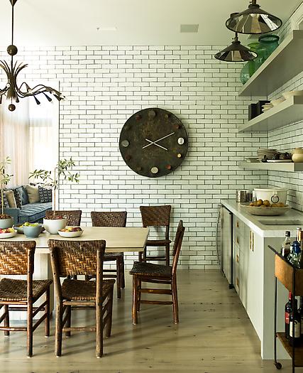 Eat in kitchen with subway tile backsplash and walls with dark grout, long wood table, reclaimed wood seats with wicker backs and seats, a chandelier, floating shelves, pendent lights and a large clock