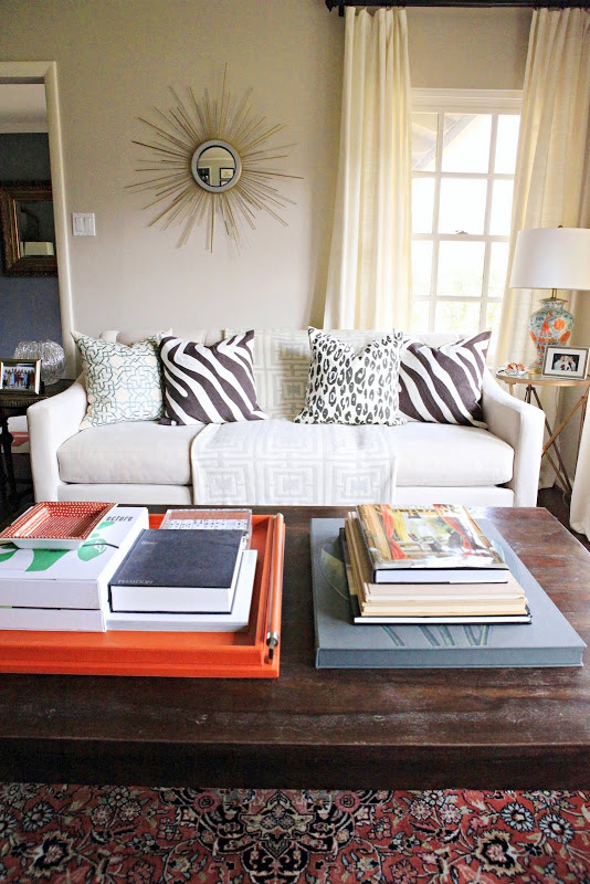 Living room with COCOCOZY Logo Throw in grey on a grey sofa with zebra and white leopard print accent pillows, a sunburst mirror, a long wood coffee table with an orange trey full of books and magazines on a Turkish rug