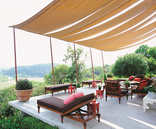 Canopy style shades cover an outdoor terrace that overlooks a hillside. Under the canopy are Moroccan style side tables and wood outdoor furniture