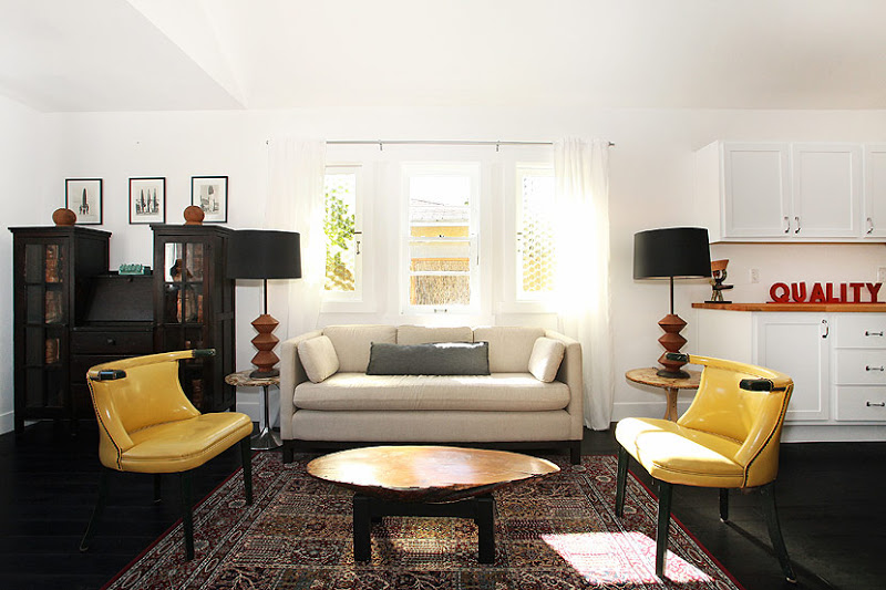 Living room in a cottage with dark wood floor, yellow settees, a taupe armchair, a painted black chest and a Turkish rug