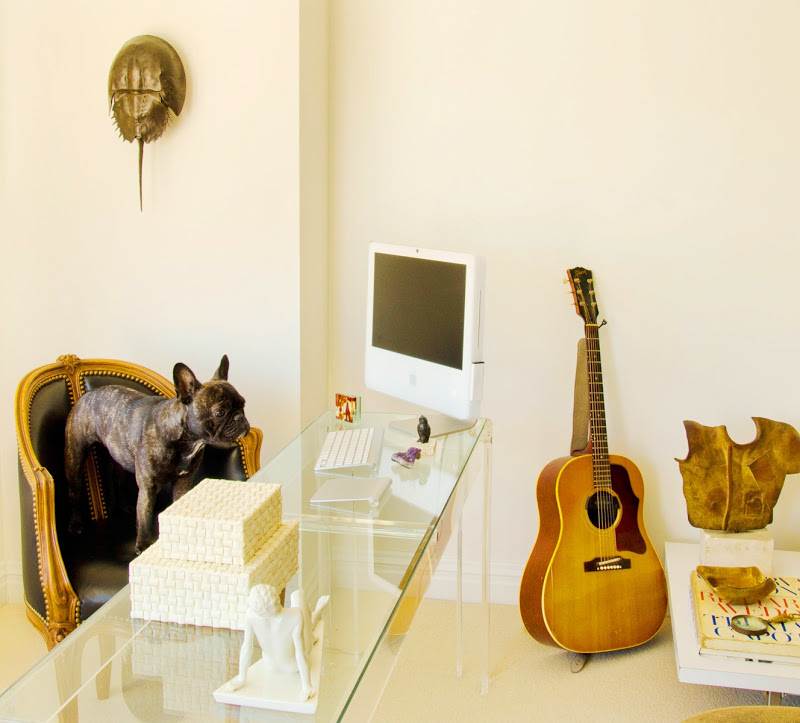 Home office with a black Louis XIV chair, a glass desk with small sculptures and a Mac computer, a guitar, a white coffee table and a black French bulldog