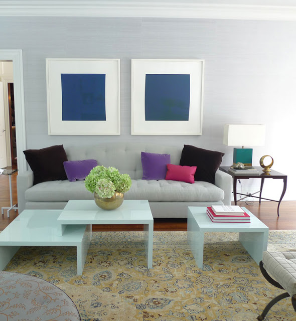 frank roop color blocked living room with purple, pink and brown pillows on a neutral sofa, two framed blue squares hang above it in a white room with wood floors, white coffee tables and yellow patterned rug