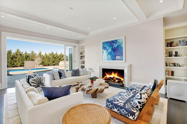 living room white high ceilings and walls, a large window overlooking the pool, a fireplace, built in bookshelf, dark wood floor, a natural trunk coffee table, two sofas and two outdoor wooden arm chairs with blue and white cushions 