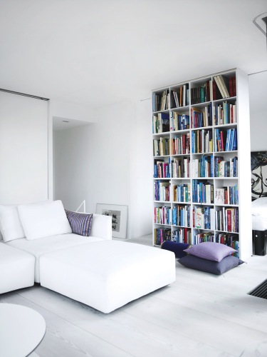 Floor to ceiling bookshelf stuffed with books dividing a small, white room with purple pillows in two