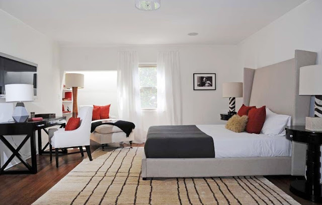 Guest bedroom with taupe wing back headboard, a black writing desk with a white armchair with red and brown accent pillows, wood floor with a striped area rug, black oval nightstands and a beige chaise longue with orange accent pillows near the window