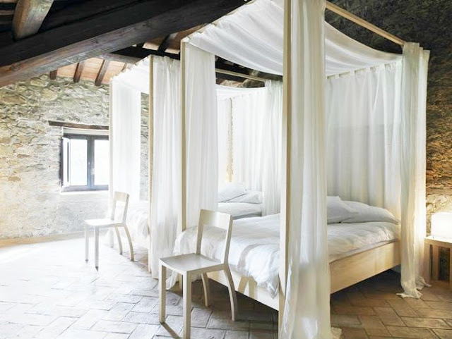 Two canopy beds in a bedroom with stone walls and floor with wispy white drapes and white linens