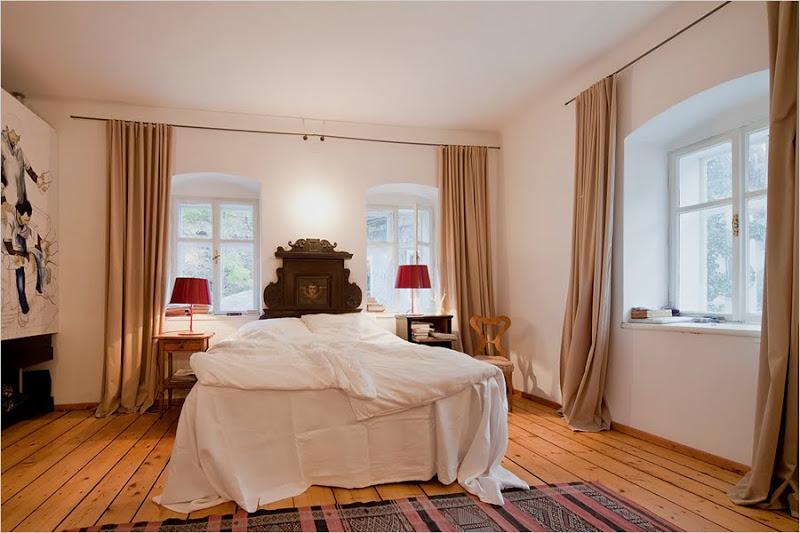 Bedroom in the restored Hammerhaus castle with knotted wood floor, large windows, floor length taupe curtains, beautifully carved wood headboard and two wood sidetables with red reading lamps