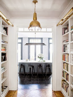 View into a NYC kitchen from a hallway with built in bookshelves, herringbone wood floors and a brass pendant light