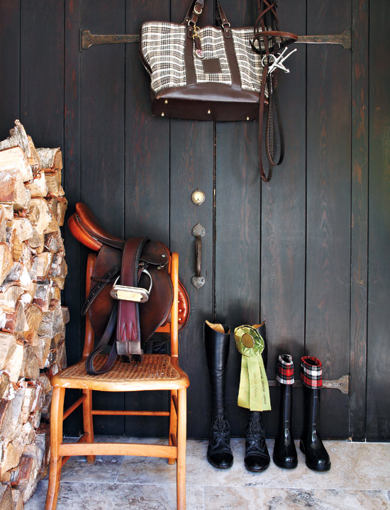 Foyer with dark wood paneled walls, a saddle draped over a wood cane chair, two pairs of riding boots, a tote bag, and a stack of firewood