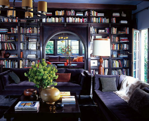 roman and williams' purple living room and den with built in bookshelves full of books bookcase, two purple sofas, a dark wood coffee table and round chandelier with yellow lampshades.