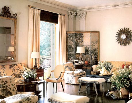 Vintage 1960s living room with chintz sofas and armchairs. The room has a dark wood floor, moulded crown detailing and a large window with floor length cream colored curtains