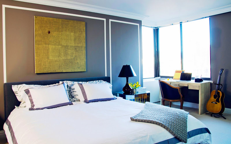 Master bedroom in Diego Garcia's NYC apartment with brown walls, an upholstered headboard, a white desk with a Louis XIV armchair and a guitar