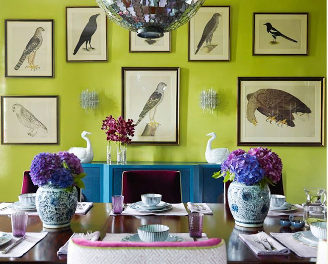 dining room in katie ridde's home with apple green wall with prints of birds, an indigo wall console table, upholstered purple chairs with gold trim, a discoball chandelier and a dark wood table with two Chinese garden pots holding purple hydrangeas