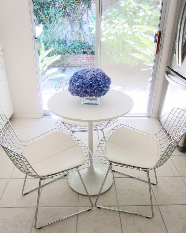 A pave arrangement of purple hydrangea on a white kitchen table surrounded by three wire chairs with white cushions