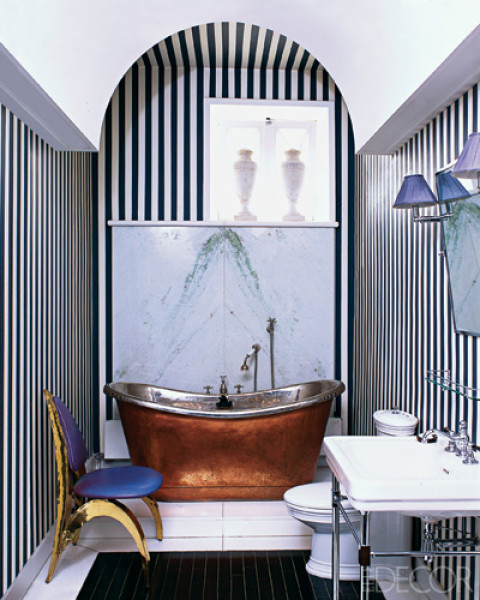 Bathroom with black and white vertical striped wallpaper, a brass stand alone tub wit ha marble backsplash, a black tile "rug" and a chair with metal legs and back painted yellow and blue cushions